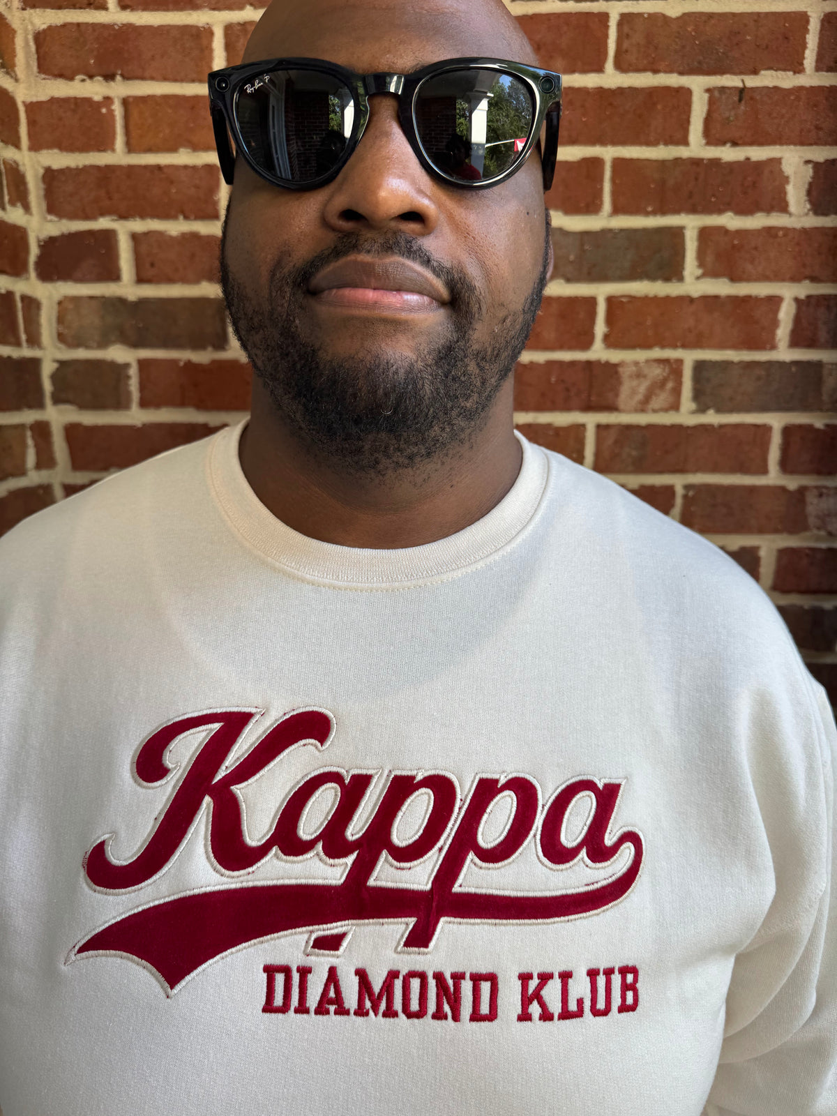 A man wearing a Kappa Alpha Psi cream crewneck sweatshirt with 'Kappa DIAMOND KLUB' printed in red across the front, standing against a red brick wall.