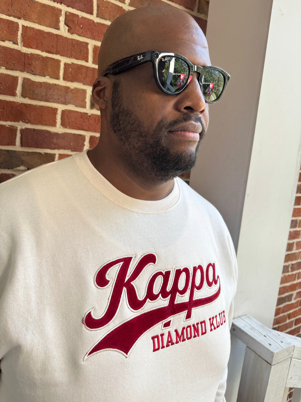 A man wearing a Kappa Alpha Psi cream-colored sweatshirt with a red 'Kappa Diamond Klue' graphic, black sunglasses, and a beard, standing in front of a red brick wall.