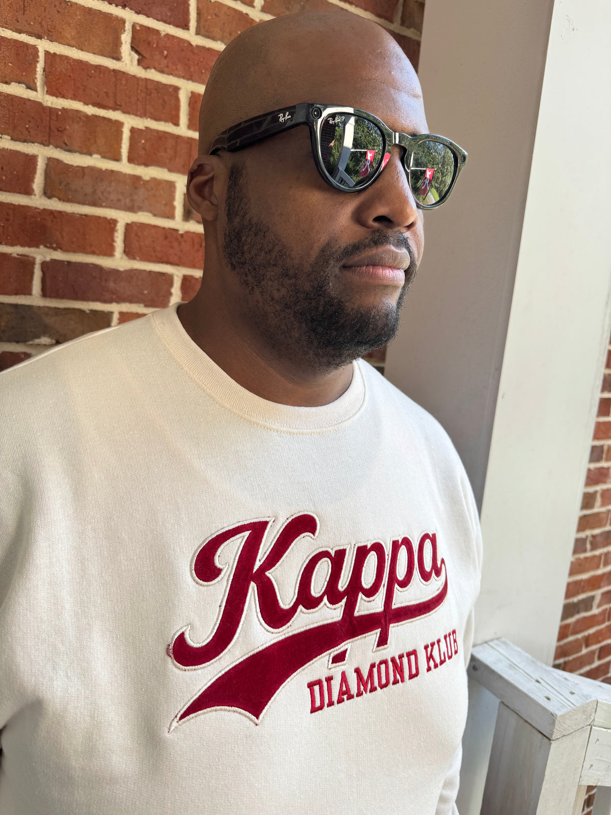 A man wearing a Kappa Alpha Psi cream-colored sweatshirt with a red 'Kappa Diamond Klue' graphic, black sunglasses, and a beard, standing in front of a red brick wall.