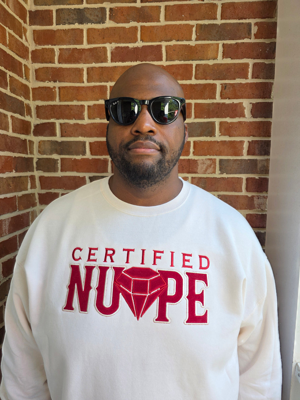 A man wearing a cream crewneck sweatshirt with a red graphic that reads 'CERTIFIED NUPE' and black sunglasses, standing in front of a red brick wall.