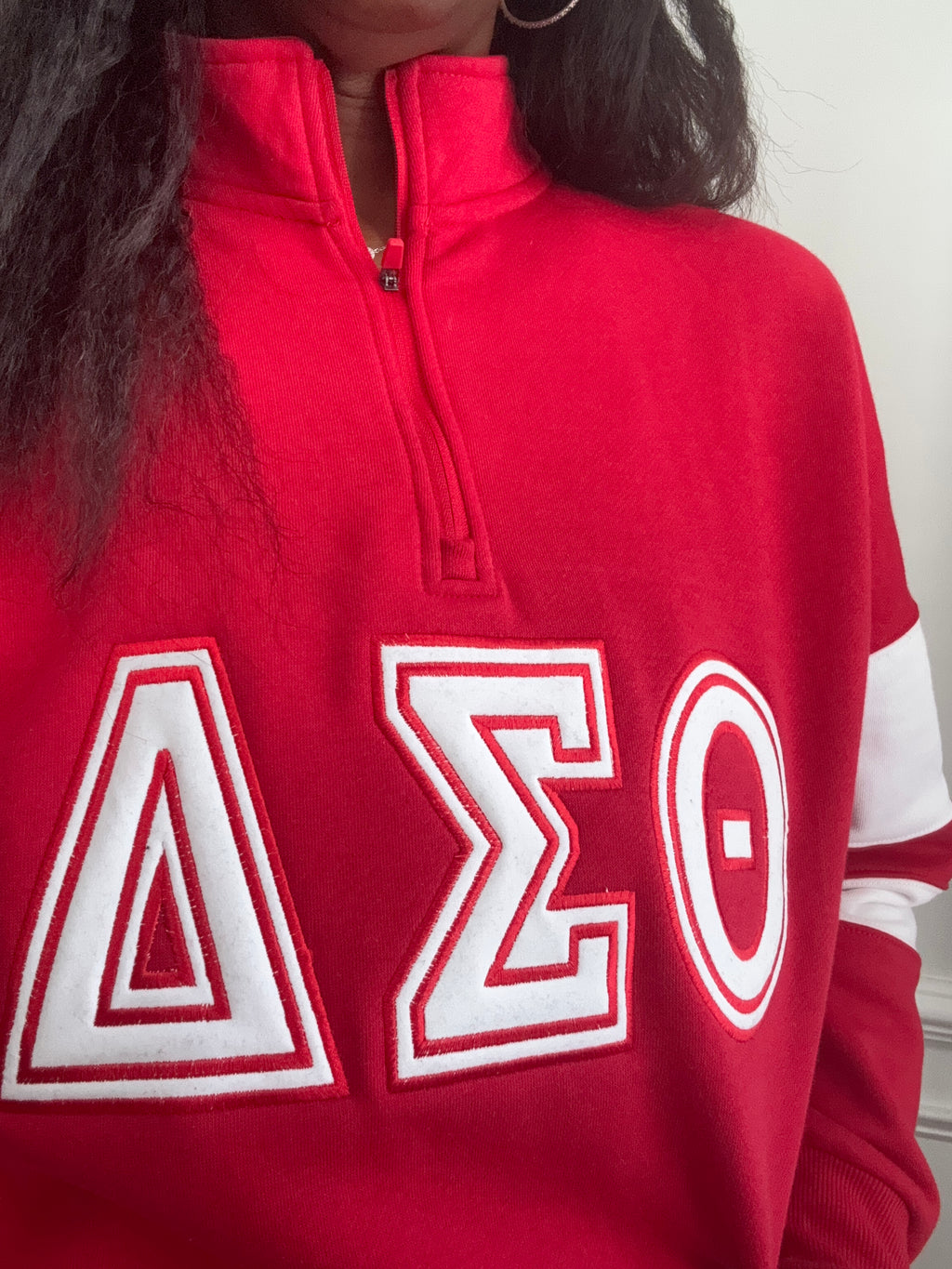 A close-up of a person wearing a red quarter-zip sweatshirt with large white embroidered Delta Sigma Theta Greek letters on the front, shown against a plain indoor background.