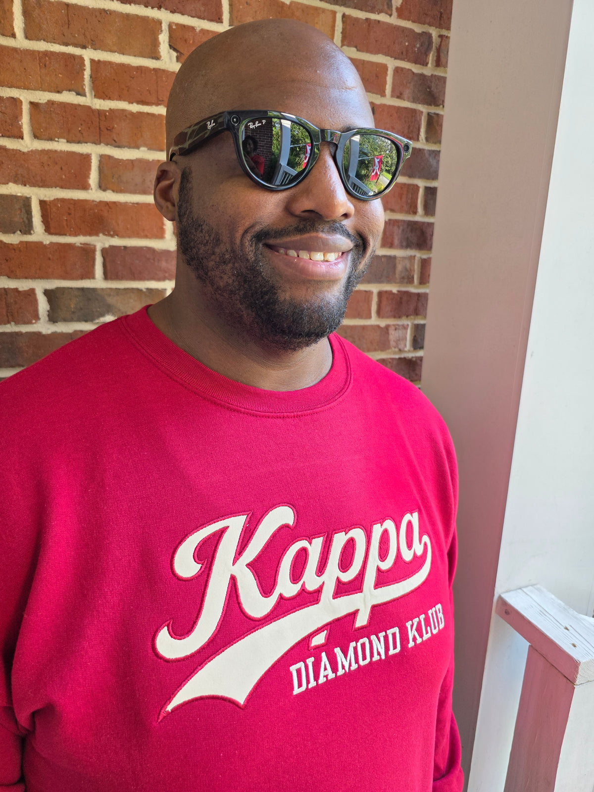 A smiling man wearing black sunglasses with mirrored lenses and a crimson Kappa Alpha Psi sweatshirt with 'Kappa DIAMOND CLUB' printed in white, standing in front of a brick wall.