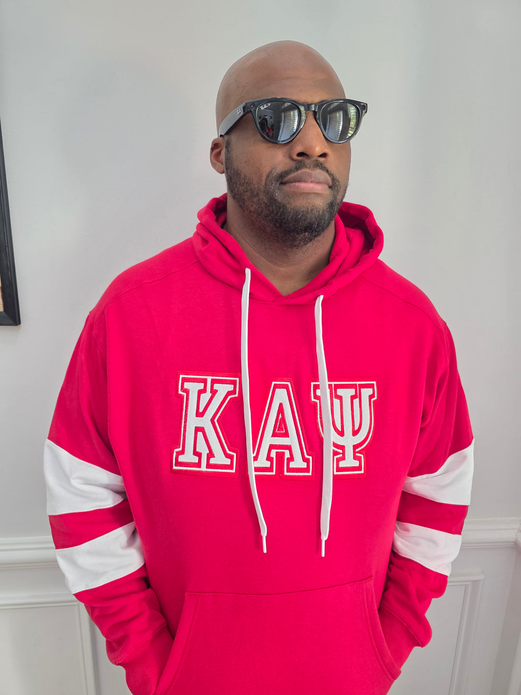A man wearing a bright red pullover hoodie with white drawstrings and white sleeve stripes, featuring the Greek letters 'KAΨ' printed in white with red outlines across the chest.