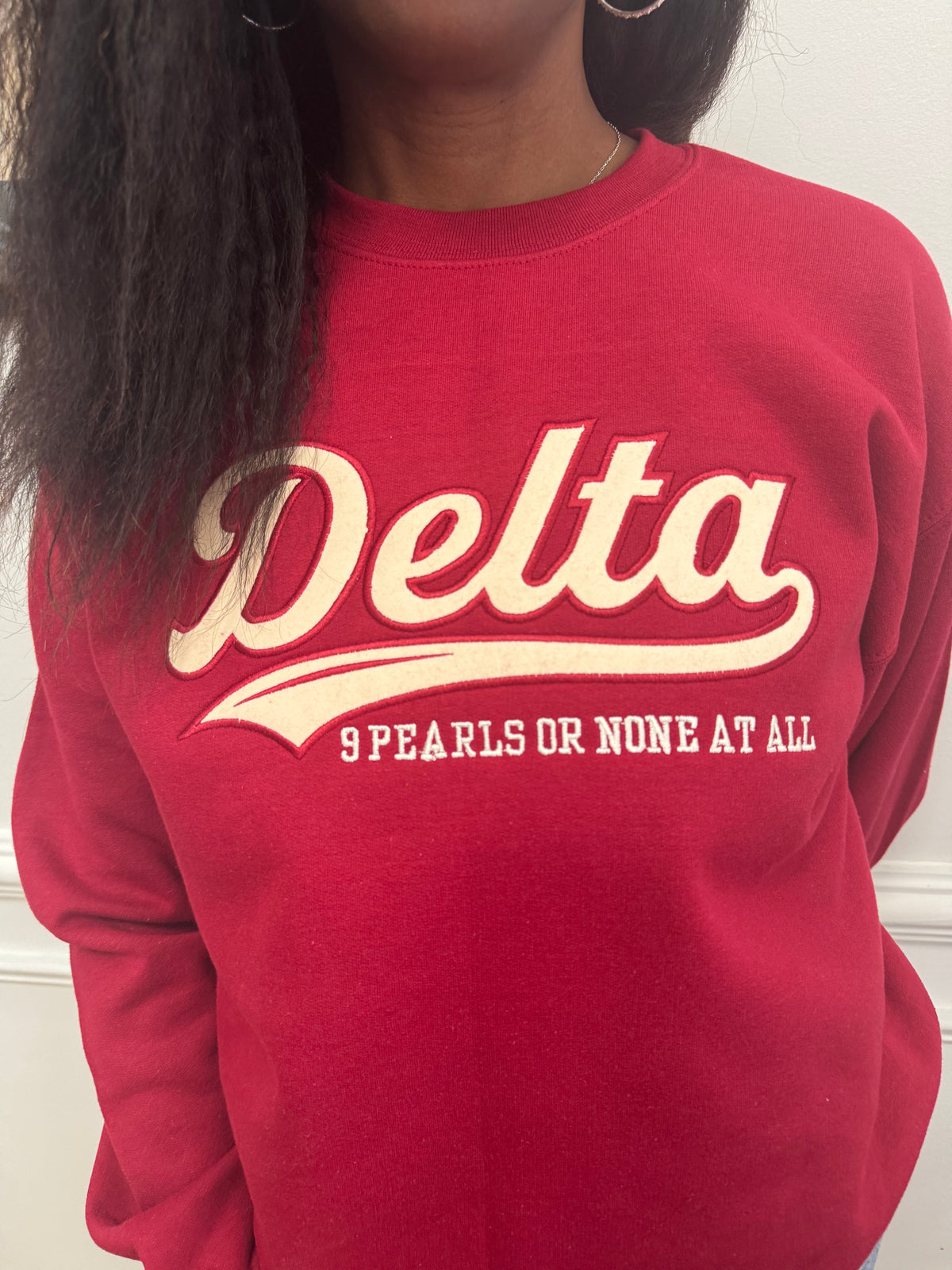 A close-up of a person wearing a red crewneck sweatshirt with a cream-colored graphic that reads 'Delta' and '9 PEARLS OR NONE AT ALL'.