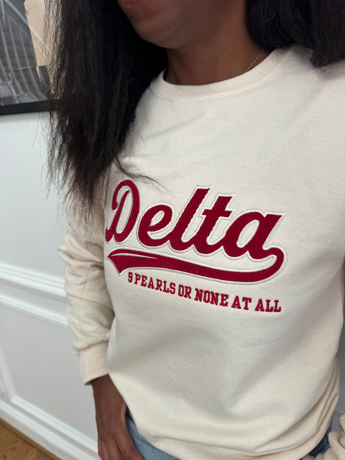 A close-up of a person wearing a cream-colored sweatshirt with a red graphic that reads 'Delta' and '9 PEARLS OR NONE AT ALL'.
